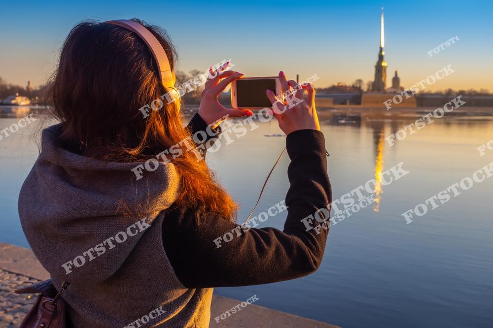 На переднем плане изображена девушка, делающая снимок Петропавловской крепости на фоне закатного неба. Золотой шпиль собора красиво отражается в спокойной воде Невы. Закат создает теплую и умиротворяющую атмосферу, подчеркивая красоту исторической архитектуры Санкт-Петербурга. Это место — один из символов города, привлекающий жителей и туристов.